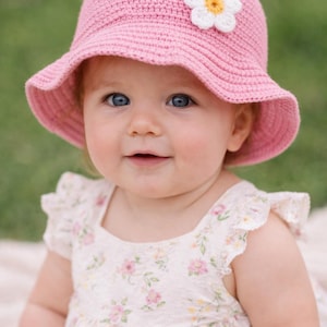 Könnte beinhalten: Ein Baby mit einem rosa gehäkelten Sonnenhut mit einer weißen und gelben Blume. Das Baby trägt auch ein weißes Kleid mit floralem Muster. Der Hintergrund ist ein verschwommenes Grün.