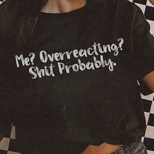 Puede incluir: Camiseta negra con el texto "Me? Overreacting? Shit Probably." La persona de la imagen lleva una gorra de béisbol a cuadros blancos y negros y gafas de sol. El fondo es un patrón de cuadros blancos y negros.