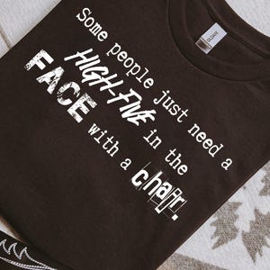 May include: Dark brown t-shirt with white text that reads "Some people just need a HIGH-FIVE in the FACE with a chair." The shirt is on a patterned surface, with a portion of a brown and white cowboy boot visible.