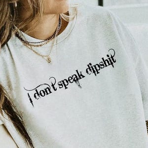 Könnte beinhalten: Hellgraues T-Shirt mit dem Text "I don't speak dipshit" in schwarzer gotischer Schrift. Das Shirt hat einen Rundhalsausschnitt und scheint aus einem weichen Material zu sein. Das Shirt wird getragen.