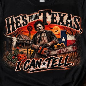 Può includere: Una t-shirt nera con una stampa grafica di una figura mascherata che tiene in mano una motosega. Il testo recita HE'S FROM TEXAS, I CAN TELL. Il design include una bandiera del Texas, una fattoria, un mulino, un teschio, un cactus e una zucca.