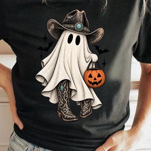 Könnte beinhalten: Ein schwarzes Kurzarm-T-Shirt mit einer Grafik eines weißen Geistes, der einen braunen Cowboyhut mit türkisfarbenem Akzent und passende Cowboystiefel trägt. Der Geist hält einen orangefarbenen Kürbiseimer, umgeben von kleinen schwarzen Fledermäusen.