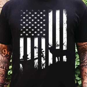 Può includere: Una t-shirt nera a maniche corte con una stampa grafica bianca della bandiera americana. Il design incorpora la sagoma di un cacciatore con arco e di un cervo nell'erba alta, integrati nel motivo delle strisce e delle stelle.