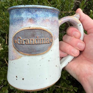 May include: A ceramic mug with a speckled white base and a blue-toned glaze at the rim. An oval-shaped plaque is centered on the front, featuring the embossed text "Grandma" in a dark brown finish with small decorative rivets on each side.