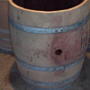 May include: A large wooden barrel with metal hoops. The barrel is made of light brown wood and has a dark brown interior. The barrel has a small hole in the side. The barrel is sitting on a concrete floor.