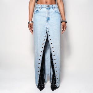 May include: Light blue denim skirt with a split front design. The skirt features metal grommets along the split edges. The skirt is paired with black boots. The skirt is a unique fashion statement.