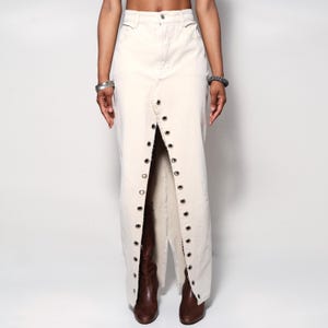 May include: Cream-colored denim skirt with a high waist and a split front. The skirt features metal grommets along the split edges. The skirt is long, reaching the ankles.