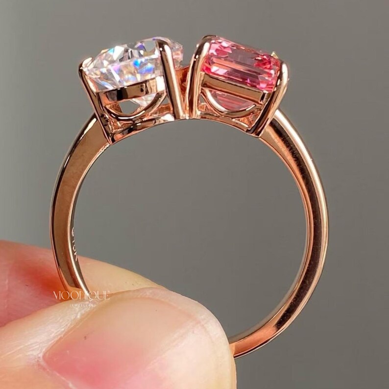 May include: Elegant Toi Et Moi engagement ring featuring a sparkling pear cut moissanite paired with a vibrant emerald cut pink sapphire in a modern two stone design. Handcrafted in solid 14K gold with a sleek polished band, this unique bridal ring symbolizes two souls united in timeless love. The text "BY MOONIQUEJEWELLERY" is visible at the bottom.