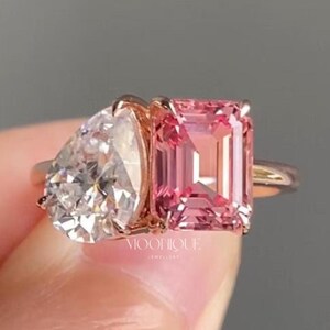 May include: Elegant Toi Et Moi engagement ring featuring a sparkling pear cut moissanite paired with a vibrant emerald cut pink sapphire in a modern two stone design. Handcrafted in solid 14K gold with a sleek polished band, this unique bridal ring symbolizes two souls united in timeless love. The text "BY MOONIQUEJEWELLERY" is visible at the bottom.