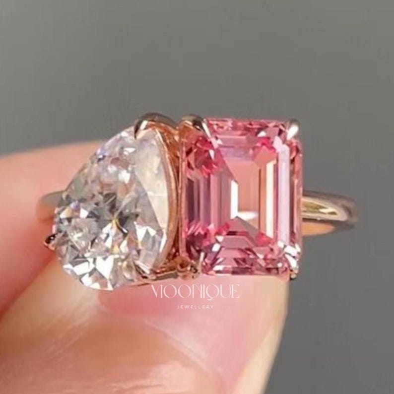 May include: Elegant Toi Et Moi engagement ring featuring a sparkling pear cut moissanite paired with a vibrant emerald cut pink sapphire in a modern two stone design. Handcrafted in solid 14K gold with a sleek polished band, this unique bridal ring symbolizes two souls united in timeless love. The text "BY MOONIQUEJEWELLERY" is visible at the bottom.