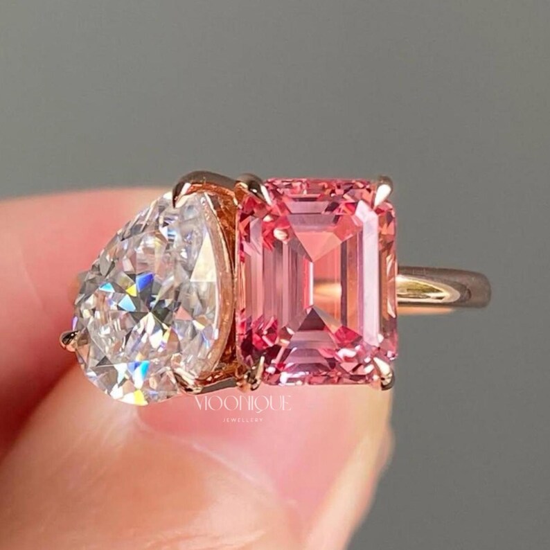 May include: Elegant Toi Et Moi engagement ring featuring a sparkling pear cut moissanite paired with a vibrant emerald cut pink sapphire in a modern two stone design. Handcrafted in solid 14K gold with a sleek polished band, this unique bridal ring symbolizes two souls united in timeless love. The text "BY MOONIQUEJEWELLERY" is visible at the bottom.