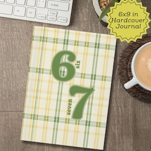 May include: A hardcover journal with a cream, yellow, and green plaid pattern. Large green numbers "6" and "7" are centered on the cover. The journal is labeled "6x9 in Hardcover Journal". A cup of coffee and a keyboard are in the background.