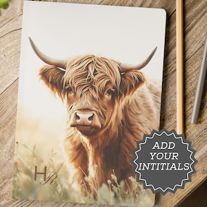 Peut inclure: Un carnet avec une illustration détaillée d'une vache des Highlands, avec une longue fourrure brune et hirsute et de grandes cornes. Le carnet affiche les initiales "HR" et le texte "ADD YOUR INITIALS". L'arrière-plan est un paysage flou.