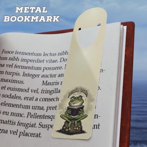 May include: A metal bookmark with a frog illustration reading a book. The bookmark is cream-colored and features the text "METAL BOOKMARK" at the top. The frog is green and sitting on a rock. The bookmark is inserted into a book.