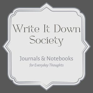 May include: A light gray framed graphic with the text "Write It Down Society" in a serif font. Below the title are the words "Journals & Notebooks" and "for Everyday Thoughts" in a sans-serif font. The background is a dark gray.