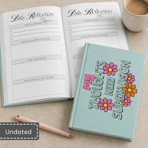 May include: A light blue journal with the text "MY THOUGHTS NEED SUPERVISON" and colourful flower illustrations on the cover. An open journal displays "Daily Reflection" pages with sections for accomplishments, gratitude, and notes. A pen is included.