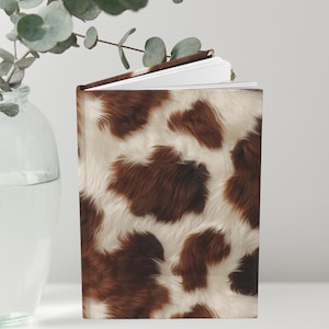 Puede incluir: Un diario de tapa dura con un patrón de piel de vaca. La cubierta presenta un diseño manchado marrón y blanco, que se asemeja al pelaje de un animal. El diario está abierto, revelando páginas blancas en el interior. Un jarrón de cristal con vegetación está en el fondo.
