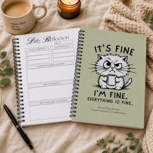 May include: A spiral-bound journal with a sage green cover featuring a grumpy cat illustration holding a mug and the text IT'S FINE I'M FINE. EVERYTHING IS FINE. The open page shows a Daily Reflection layout with sections for tasks and gratitude.