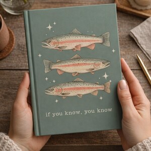 May include: A teal notebook with three fish illustrations, each with pink and silver accents, and the text "if you know, you know." The notebook is held by a person.