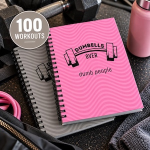 Könnte beinhalten: Zwei spiralgebundene Notizbücher mit Wellenlinienmuster. Das obere Notizbuch ist leuchtend pink mit schwarzem Text DUMBELLS OVER dumb people und einer Hantelgrafik. Das untere Notizbuch ist grau. Ein kreisförmiges Abzeichen zeigt 100 WORKOUTS.