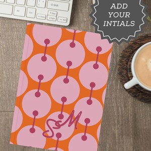 May include: A personalized notebook with a vibrant orange cover featuring a pink circular pattern and the initials "S&M". The notebook is next to a cup of coffee and a keyboard.