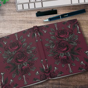 May include: A burgundy journal with a floral rose and bone design. The open journal displays a repeating pattern of dark red roses, gray leaves, and bone-shaped accents. A black pen and keyboard are visible in the background.