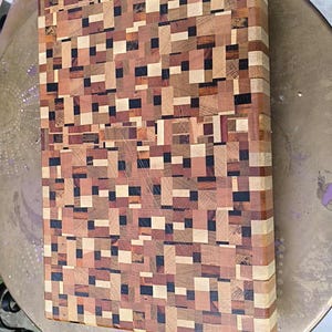 Peut inclure: Une planche à découper rectangulaire en bois avec un motif en mosaïque de différentes teintes de bois, dont des bruns clairs, moyens et foncés, et des accents noirs. La planche mesure environ 30 cm de long et 20 cm de large, posée sur une table ronde en métal.