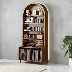 May include: A wooden arched bookcase with shelves filled with books, CDs, and vinyl records. The bookcase has a built-in turntable and speakers. A small clock displays the number "1907". A green chair and a potted plant are in the background.