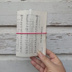 May include: A rectangular sheet of off-white paper featuring black printed musical notation and lyrics for the hymn It Is Well with My Soul. The paper is secured horizontally by a thin, bright pink elastic band across the center of the page.