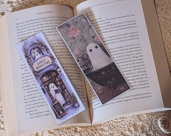May include: Two rectangular paper bookmarks resting on an open book. One bookmark features a ghost in a bookstore with the text GHOST BOOKSTORE and NEW SPIRITS DAILY. The other bookmark shows a ghost in a bathtub surrounded by pink roses.