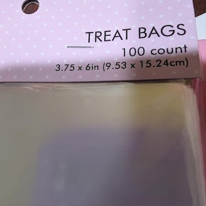 以下が含まれることがあります： 白い水玉模様のライトピンクのヘッダーが付いた、100枚入りの透明なトリートバッグ。パッケージには「TREAT BAGS」「100 count」「3.75 x 6in (9.53 x 15.24cm)」と記載されています。バッグは透明で、パッケージ内に重ねて収納されています。