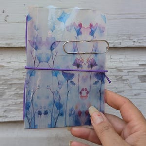 May include: A small, translucent notebook cover featuring a repeating watercolor-style floral pattern in shades of blue, purple, and pink. The cover is secured with a thin purple elastic band and includes a single silver metal paper clip attached.
