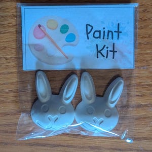 以下が含まれることがあります： 透明なビニール袋に入った2つの白いウサギ型のフィギュアと、Paint Kitという文字と絵筆付きのパレットのイラストが描かれた紙のラベル。ウサギのフィギュアには、成形された耳、丸い目、ハート型の鼻が付いています。