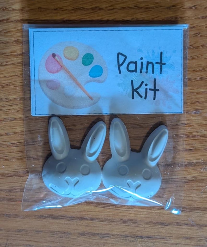 以下が含まれることがあります： 透明なビニール袋に入った2つの白いウサギ型のフィギュアと、Paint Kitという文字と絵筆付きのパレットのイラストが描かれた紙のラベル。ウサギのフィギュアには、成形された耳、丸い目、ハート型の鼻が付いています。