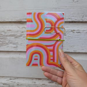 May include: A small rectangular notebook featuring a vibrant pattern of wavy, concentric lines in shades of orange, pink, red, and light blue. The notebook is secured with a bright lime green elastic band and includes a gold-toned metal paper clip.