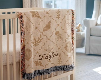 May include: A woven throw blanket draped over a crib, featuring a tan background with a repeating pattern of brown seashells, starfish, coral, and jellyfish. The name Taylor is embroidered in brown script, and the edges have a multicolored fringe.