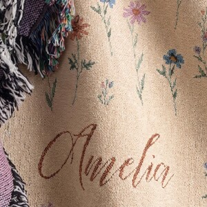 May include: A woven throw blanket in a light beige color featuring a pattern of small blue, pink, and orange wildflowers. The name Amelia is embroidered in a brown cursive script. The blanket has a textured fringe edge with purple and black threads.