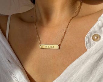 May include: A gold-toned metal bar necklace featuring a delicate chain and a rectangular pendant engraved with the lowercase word mama in a simple sans-serif font, resting against a white collared shirt with a visible button.