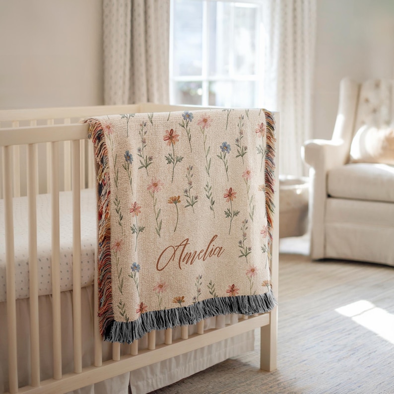 May include: A woven throw blanket featuring a cream-colored base with a repeating pattern of small, colorful wildflowers in shades of pink, blue, and orange. The name Amelia is embroidered in a brown script font, and the edges have a fringed trim.