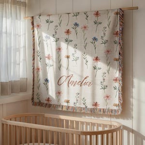 May include: A woven throw blanket featuring a repeating pattern of watercolor-style wildflowers in shades of pink, orange, and blue on a cream background. The name Amelia is printed in a brown cursive script. The blanket has fringed edges.