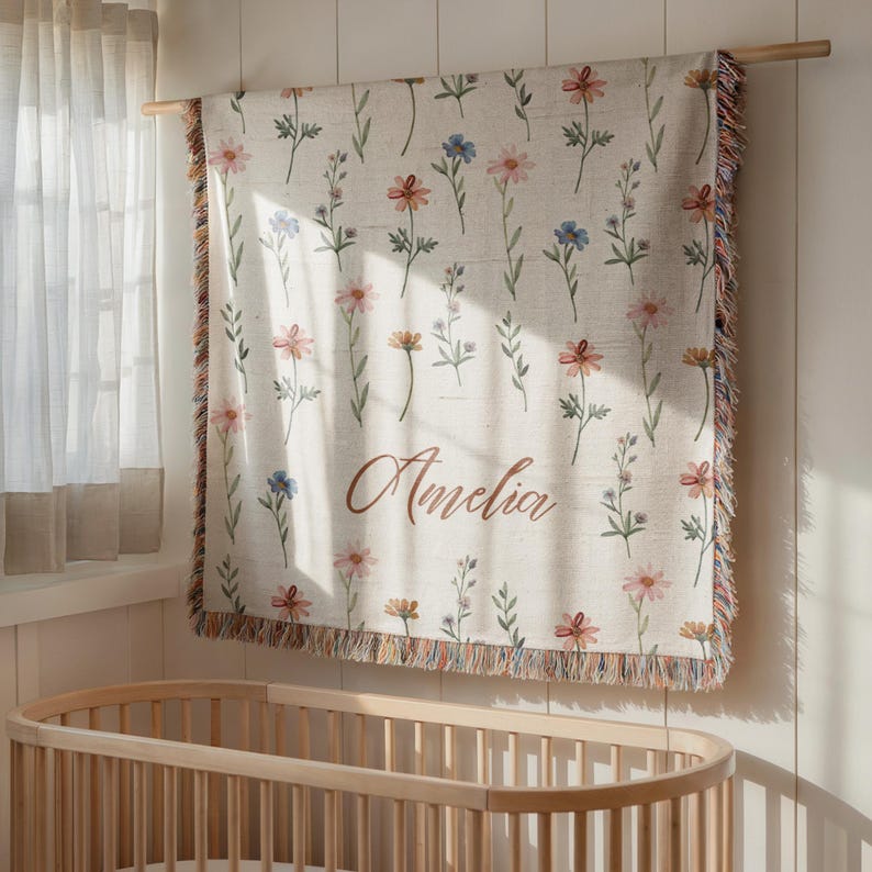 May include: A woven throw blanket featuring a repeating pattern of watercolor-style wildflowers in shades of pink, orange, and blue on a cream background. The name Amelia is printed in a brown cursive script. The blanket has fringed edges.
