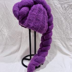 May include: A purple knit hat with a braided design. The hat is a vibrant shade of purple and features a ribbed knit pattern on the main part of the hat. The braid extends down from the back of the hat.