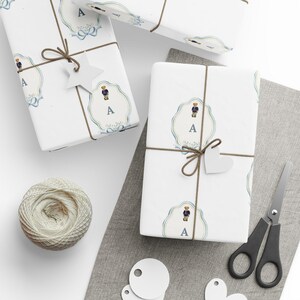 Peut inclure: Plusieurs boîtes cadeaux blanches, décorées d'un motif répété d'un ours en peluche en blazer bleu marine et de la lettre "A". Les cadeaux sont attachés avec de la ficelle marron et ornés d'étiquettes en papier blanc. Une pelote de ficelle et des ciseaux sont également visibles.