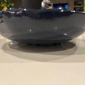 May include: A navy blue ceramic bowl with a wavy rim. The bowl has a glossy finish and sits on a white surface. The bowl's shape is round and shallow, suitable for serving or display.