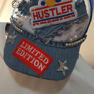 Puede incluir: Gorra de camionero de mezclilla con una parte trasera de malla blanca y un borde azul. La gorra presenta la palabra "HUSTLER" en un parche rojo y blanco, junto con el texto "GETTIN' BREAD AND MAKIN' MY DOUGH RISE". También tiene un parche de "EDICIÓN LIMITADA".