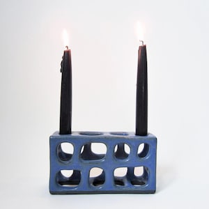 Può includere: Un portacandele in ceramica blu con un design rettangolare, caratterizzato da molteplici aperture quadrate. Due candele nere sono accese, poste nel supporto. Il portacandele è un oggetto decorativo per l'arredamento della casa.