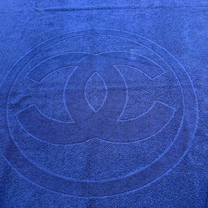 May include: A blue beach towel with a large, embossed logo in the center. The logo is a double-C design within a circular border. The towel is made of a soft, absorbent material, perfect for use at the beach or pool.
