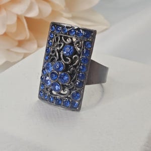 May include: A rectangular ring with a dark metal setting. The ring features a central design of blue gemstones surrounded by a filigree pattern and additional blue stones along the edges. The ring is set against a soft, neutral background.