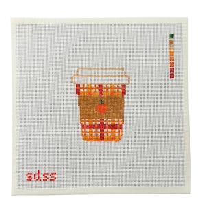 May include: A needlepoint design featuring a coffee cup with a plaid pattern in orange, red, and yellow. The cup has a brown sleeve with an orange apple. The letters "sdss" are in red at the bottom left.