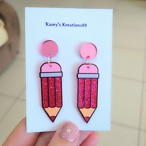 May include: A pair of dangle earrings shaped like pencils on a white display card labelled Kasey's Kreations88. The earrings feature a pink circular stud, a light pink eraser, a grey band, and a body filled with red glitter, ending in a peach tip.